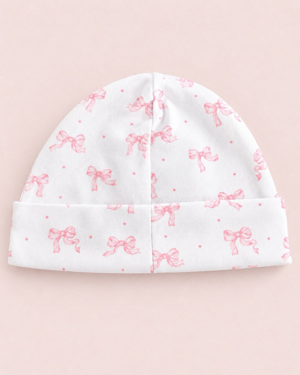 A white knit baby beanie hat with a pink bow and dot pattern, folded at the brim, shown against a solid pale pink background.