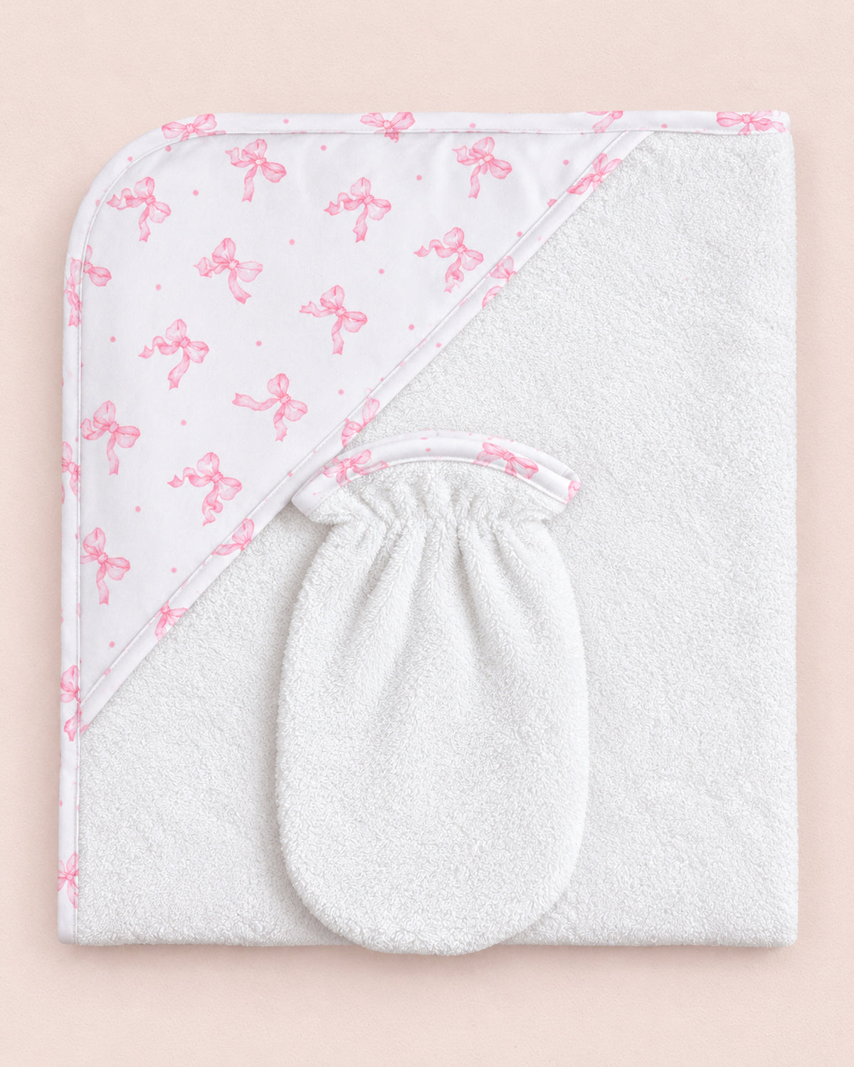 A white hooded baby towel with a soft textured body and a patterned hood panel featuring pink bows and small pink dots, laid flat on a pale pink background.