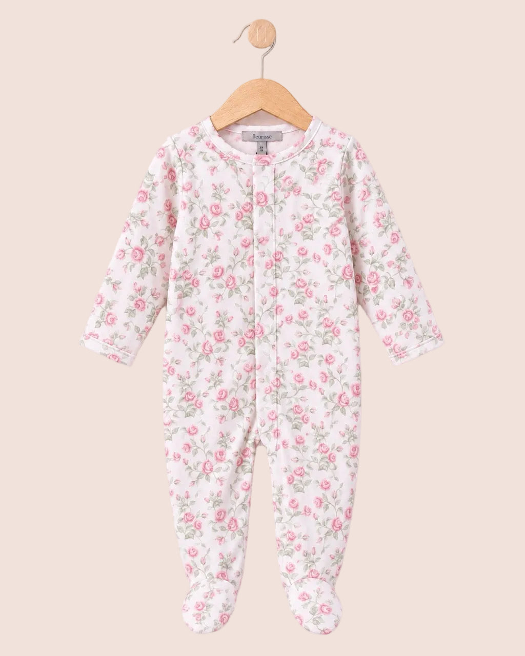 A baby footed one-piece sleeper with a pink and green floral print on a white background, hanging on a wooden hanger against a pale pink background.