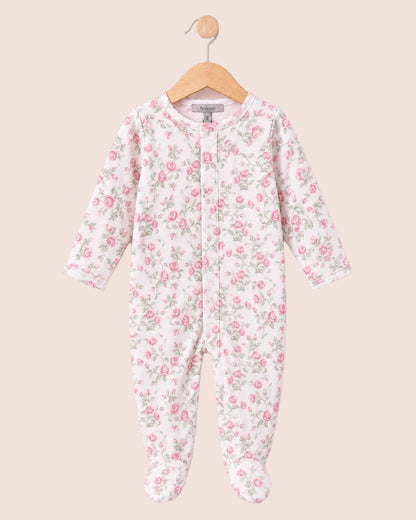 A baby footed one-piece sleeper with a pink and green floral print on a white background, hanging on a wooden hanger against a pale pink background.