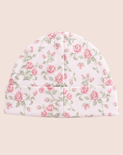 A soft fabric beanie hat with a pink rose and green leaf floral pattern on a white background, laid flat against a solid pale pink backdrop.