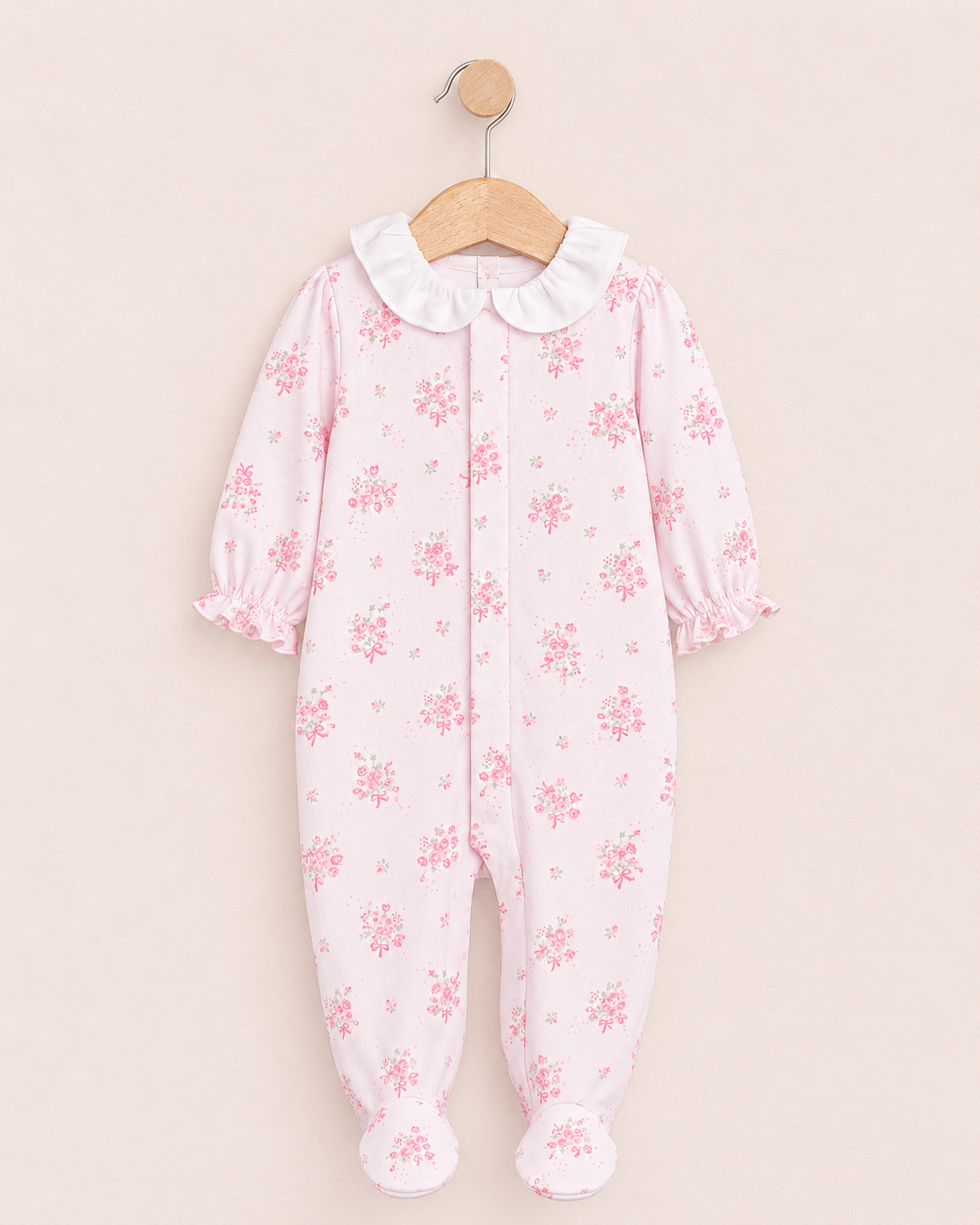 Baby stretchie hanging from wooden hanger with white collar and light pink textured fabric featuring a delicate floral print with pink roses, green leaves, and pink ribbon bows