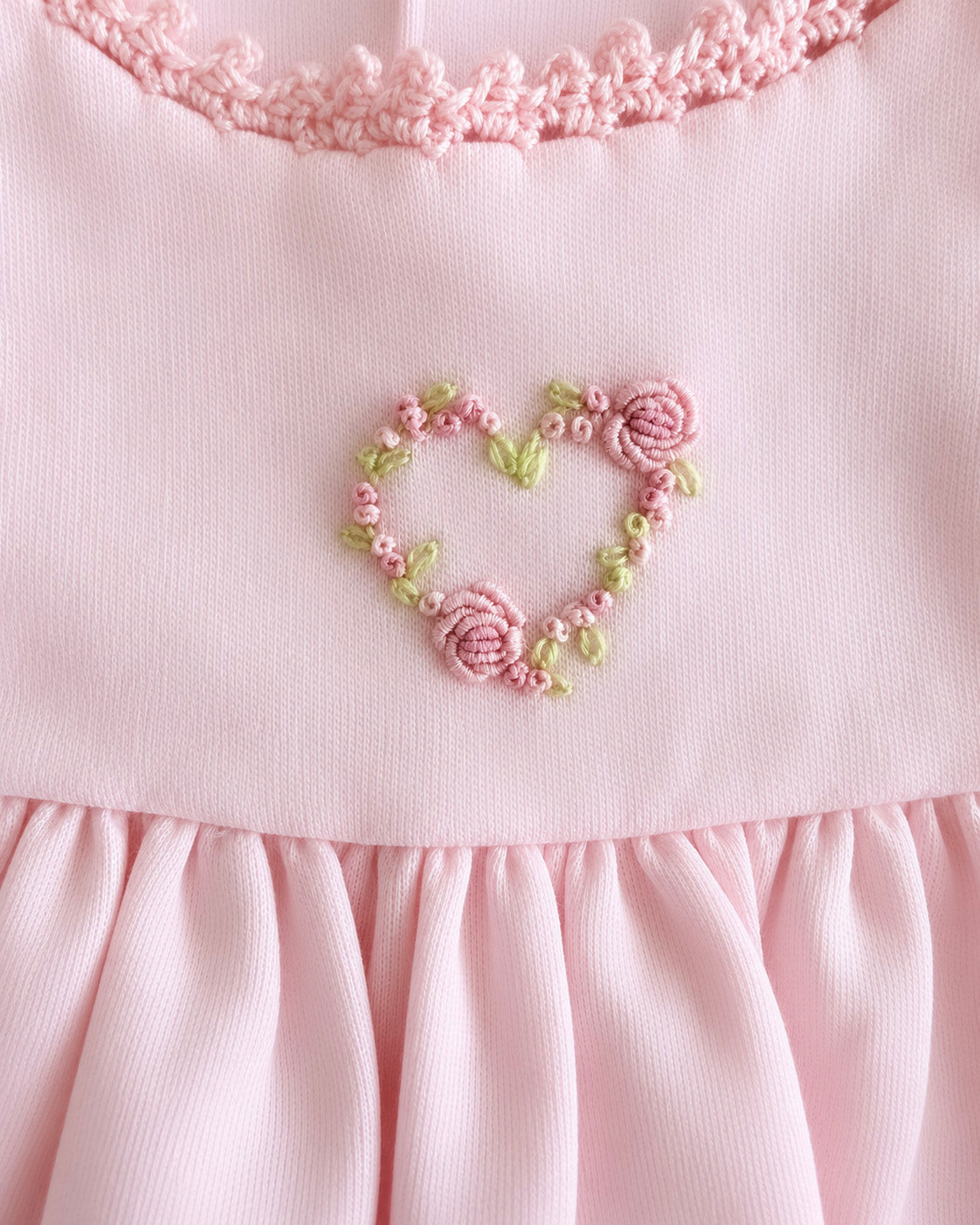 Close-up of a light pink dress featuring a heart-shaped floral embroidery in pink and green thread, with a ruffled neckline and pleated skirt.