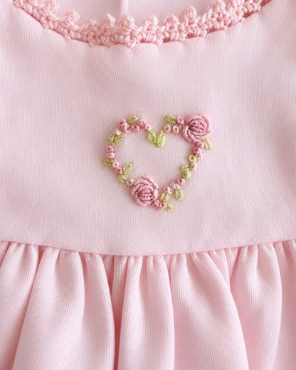 Close-up of a light pink dress featuring a heart-shaped floral embroidery in pink and green thread, with a ruffled neckline and pleated skirt.