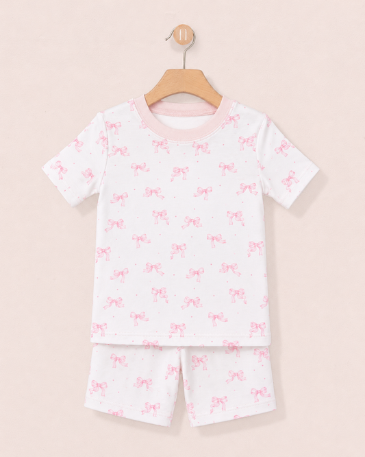 A 2-piece pajama set with short sleeves and short bottoms, in white, with a pink bow and dot pattern, hanging on a wooden hanger against a solid pale pink background.
