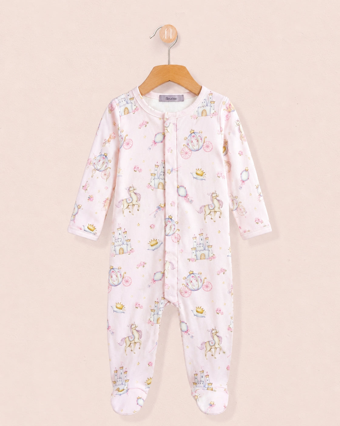 A baby one-piece stretchie hanging on a wooden hanger against a light beige background, in a pale pink color, decorated with a whimsical print of unicorns, castles, and bicycles in pastel blue, yellow, and green.