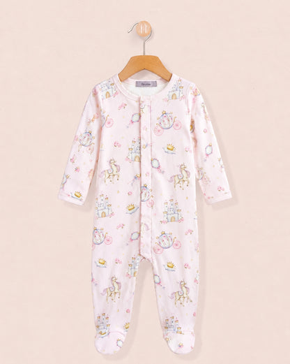 A baby one-piece stretchie hanging on a wooden hanger against a light beige background, in a pale pink color, decorated with a whimsical print of unicorns, castles, and bicycles in pastel blue, yellow, and green.