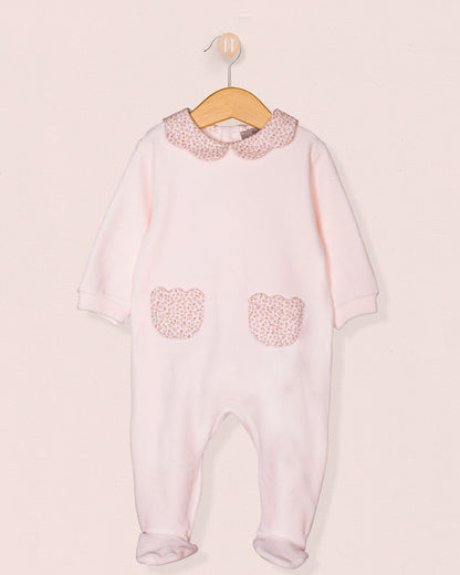 Full front view of a baby girl's pink footie romper made from soft Italian velour, featuring long sleeves, a floral scalloped collar, and two heart-shaped front pockets in a matching floral print. The romper is displayed on a wooden hanger against a soft blush background.