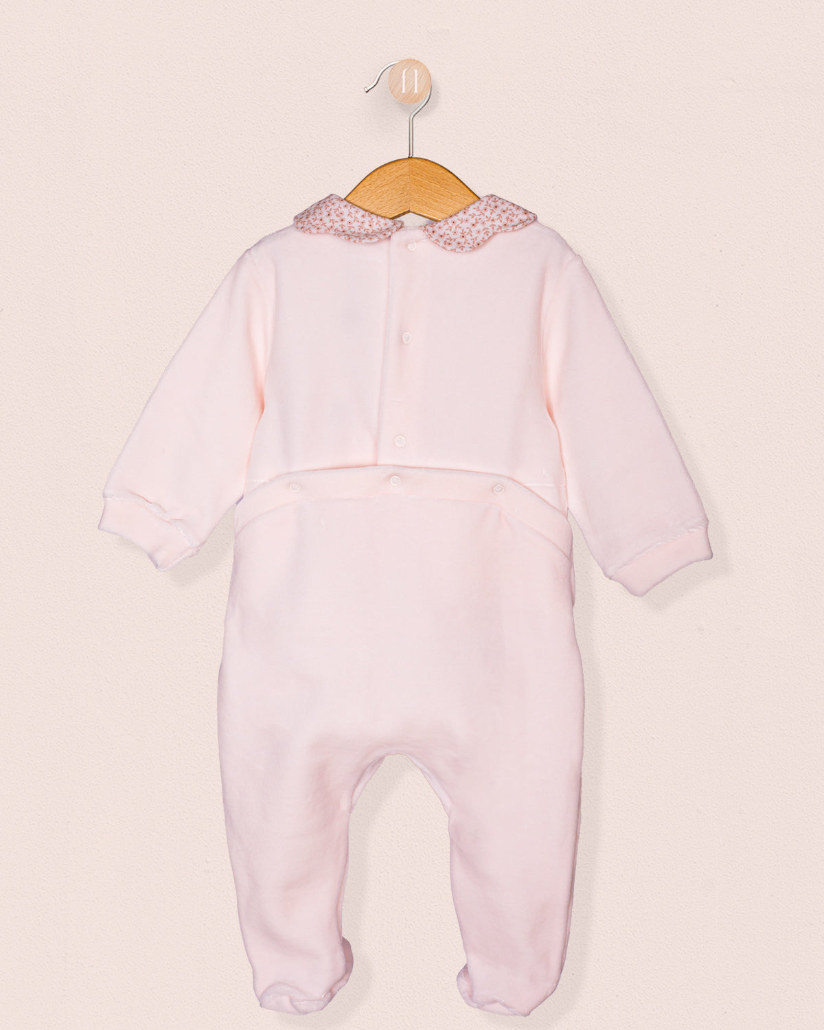 Back view of the baby girl's pink footie romper showing the full-length design with a floral collar and concealed snap closures along the back for easy dressing, displayed on a wooden hanger against a blush-toned backdrop.