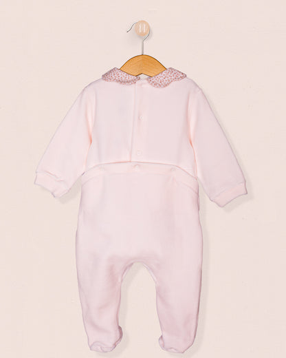 Back view of the baby girl's pink footie romper showing the full-length design with a floral collar and concealed snap closures along the back for easy dressing, displayed on a wooden hanger against a blush-toned backdrop.