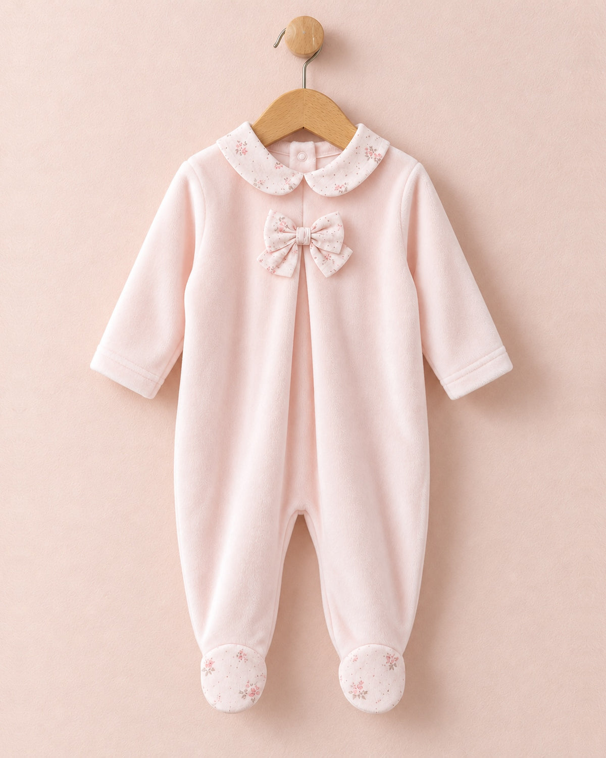 Front of light pink velour baby onesie hanging from wooden hanger, with light pink bows in center of chest, and pink floral motif collars and feet.
