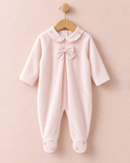 Front of light pink velour baby onesie hanging from wooden hanger, with light pink bows in center of chest, and pink floral motif collars and feet.