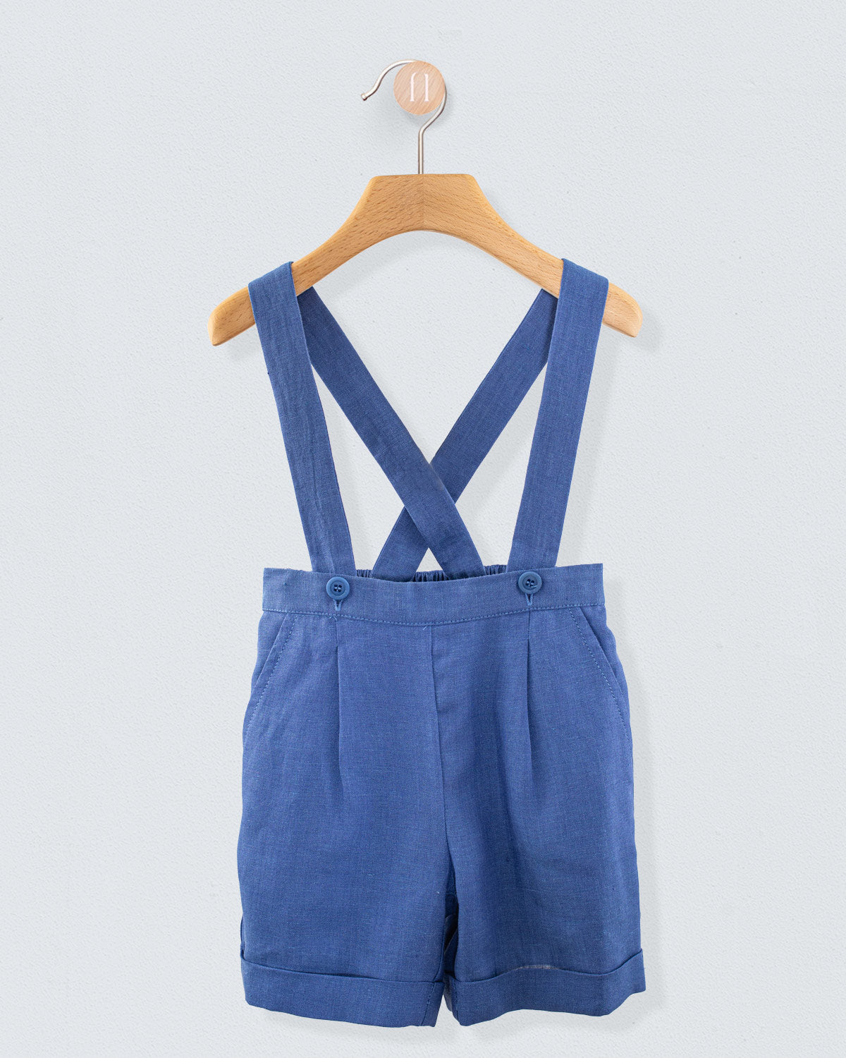 Front view of blue linen suspender shorts with crossed straps and cuffed hem, displayed on a wooden hangert