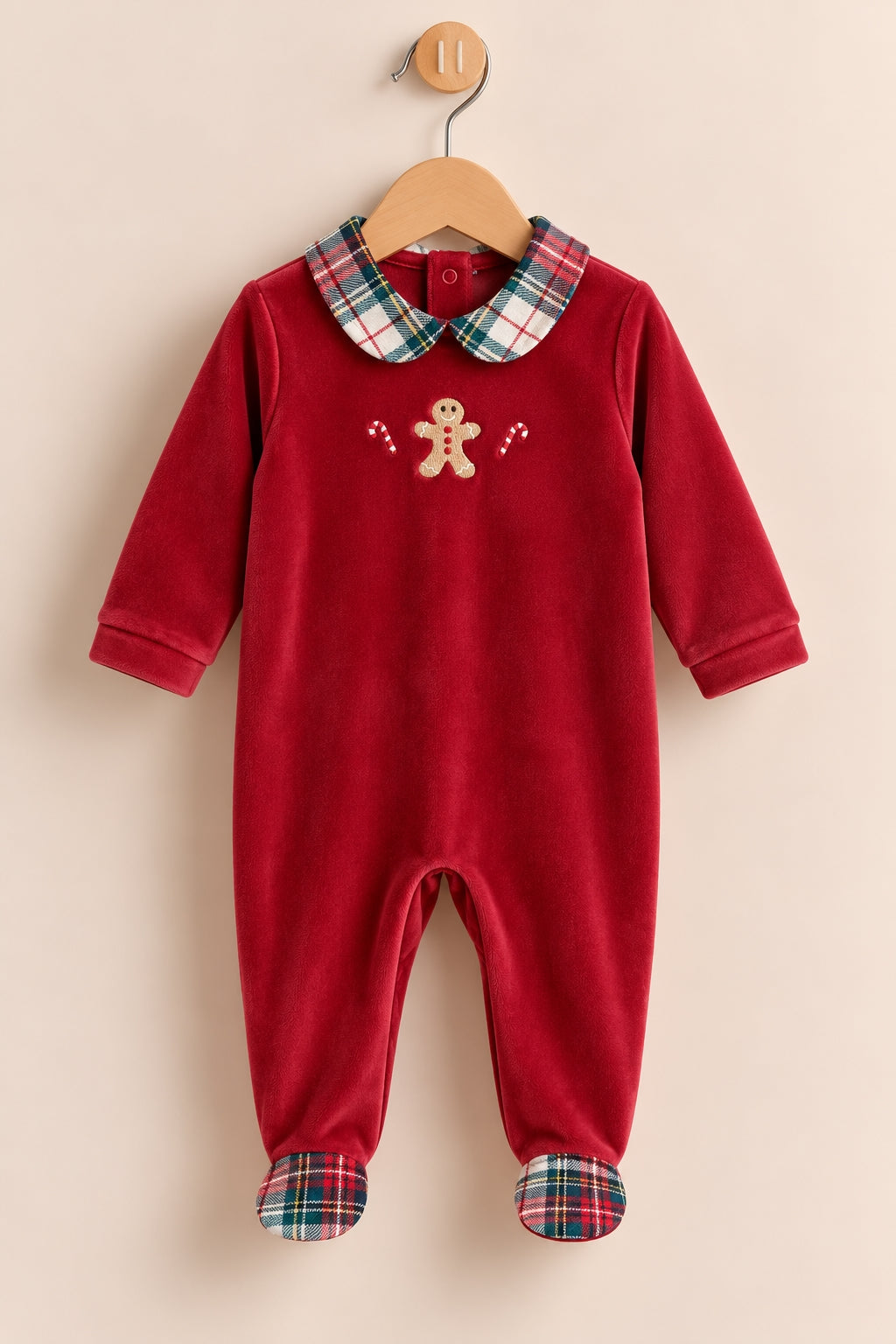 A baby footed one-piece jumpsuit in deep red velvet, featuring a green and red plaid collar and matching plaid cuffs, with a small gingerbread man embroidery on the chest. The garment is displayed on a wooden hanger against a solid beige background.