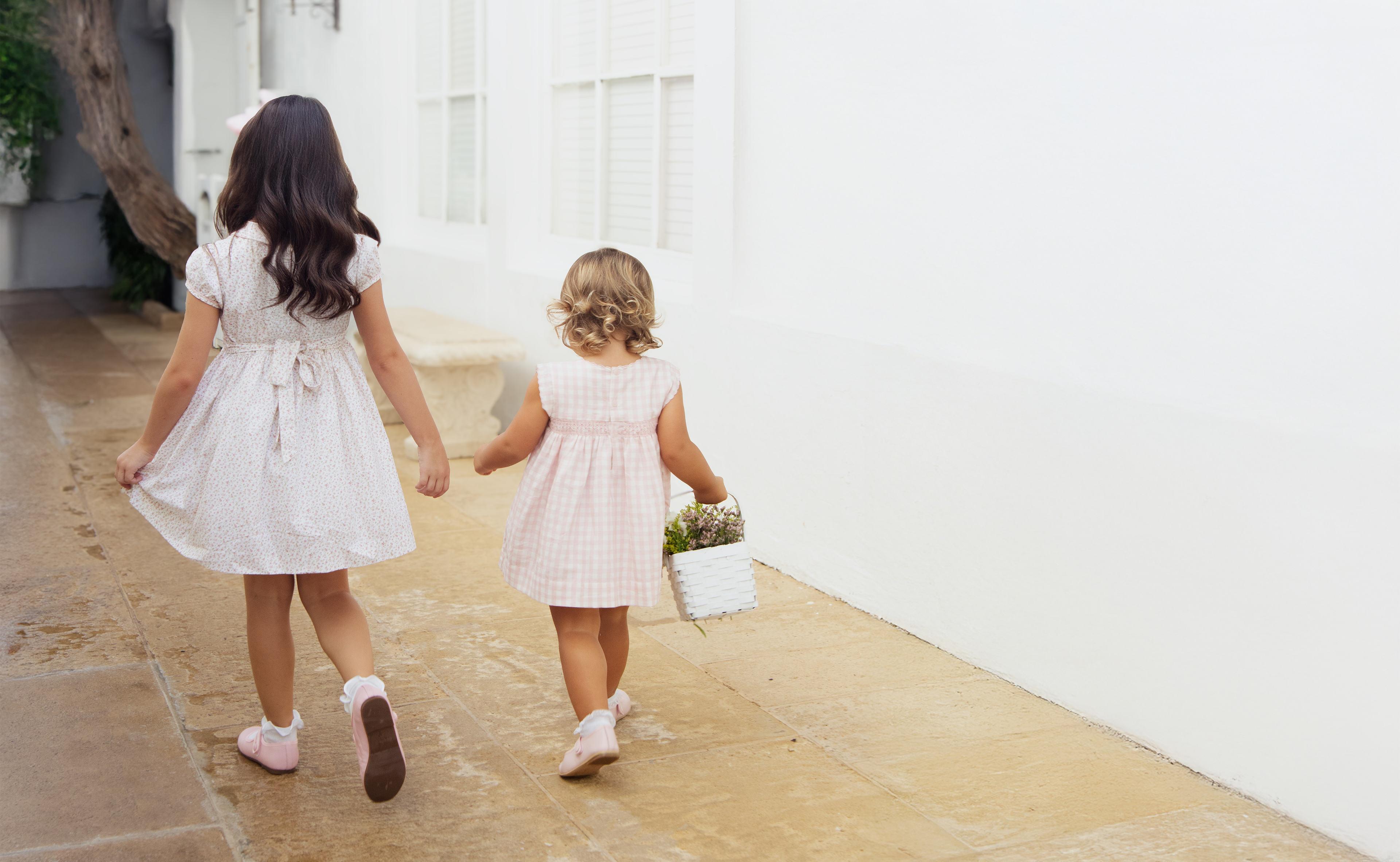 Two young girls in white dresses walking hand-in-hand on a stone path.