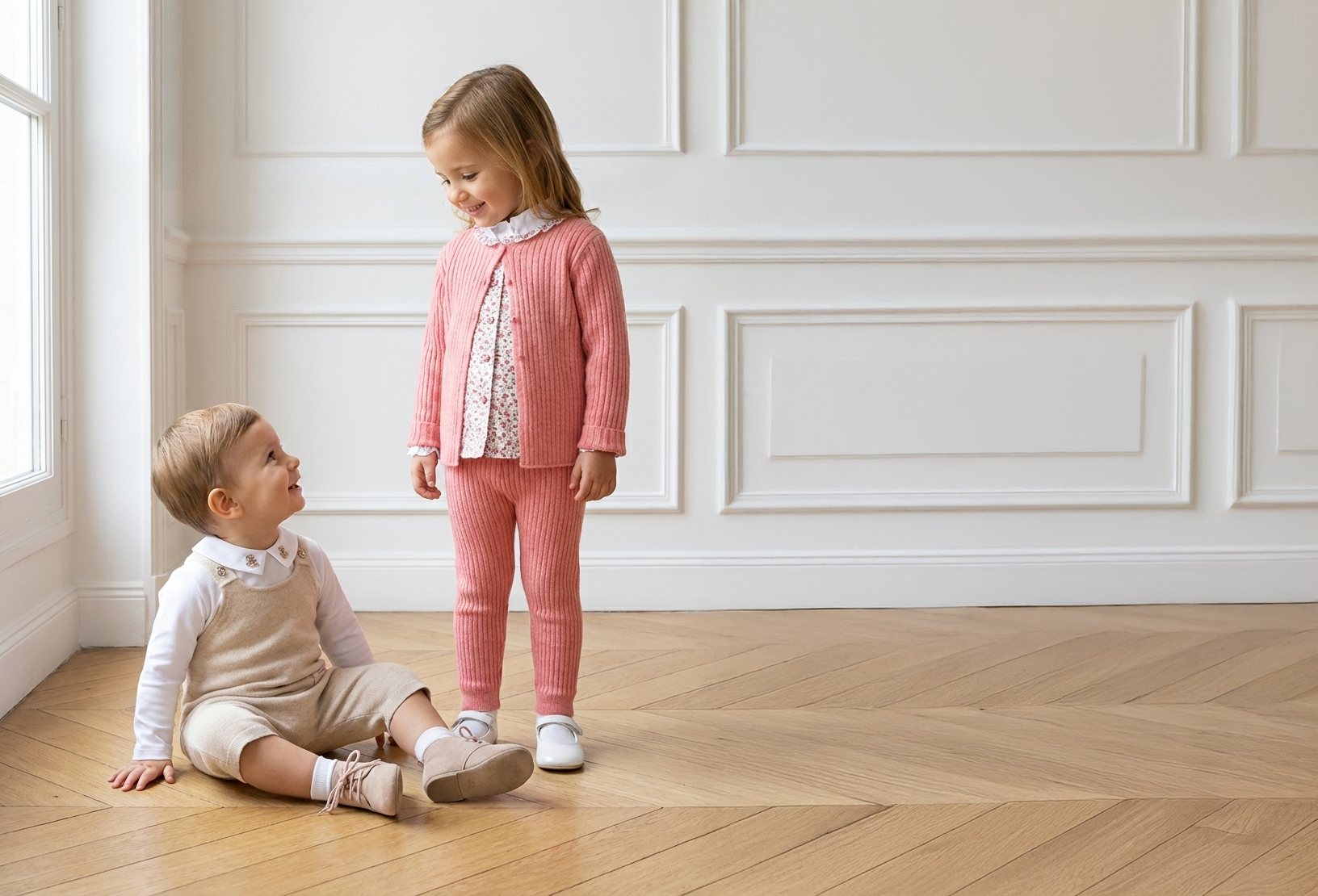 girl in pink outfit looking down at small boy looking up from the floor, on a wood floor and white wall background with french molding, by a window