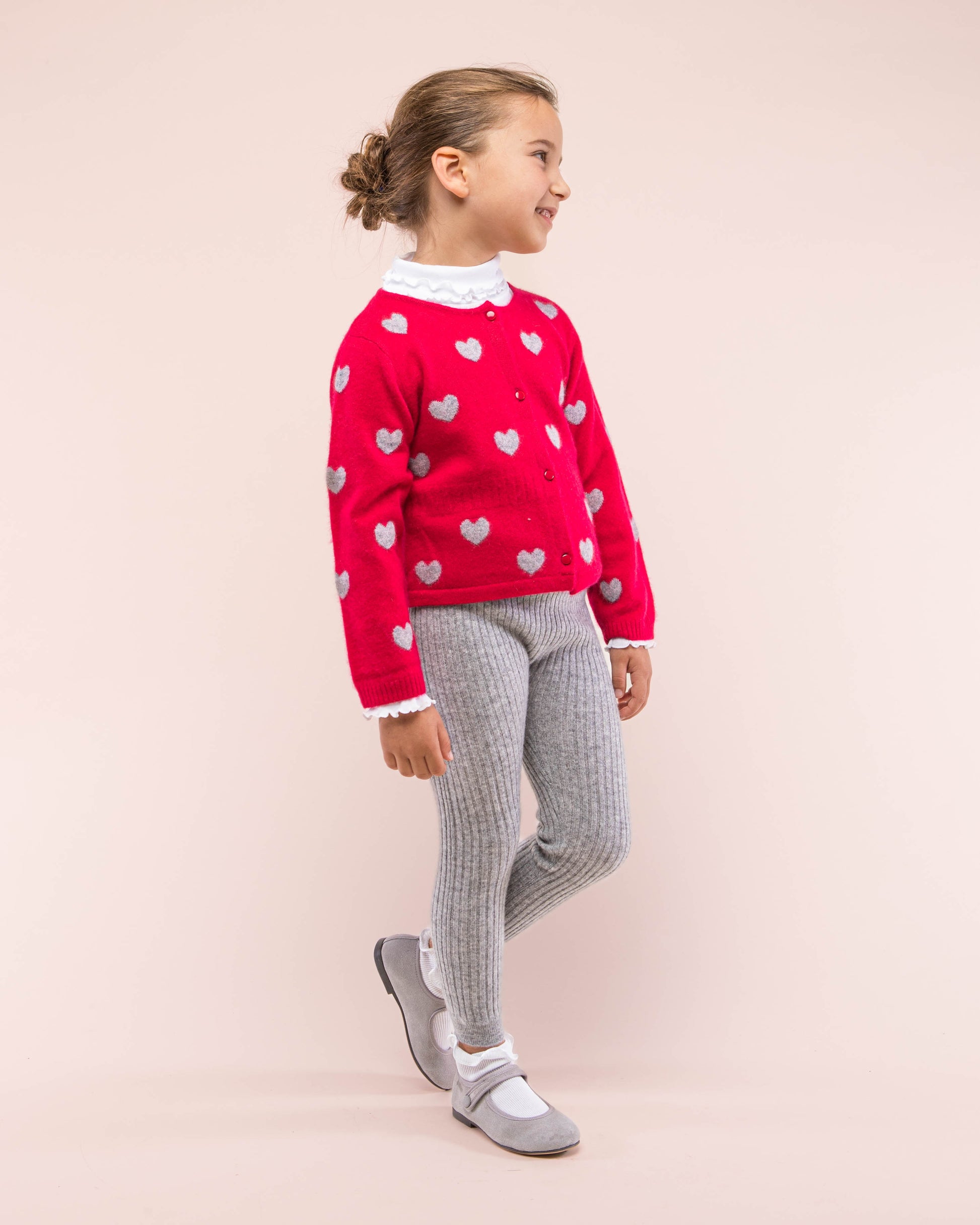 Girl model wearing Madeleine Red Hearts Cashmere sweater facing to the front looking to the side showing the front details of the sweater; paired with leggings and shoes in contrasting grey color and a white turtleneck