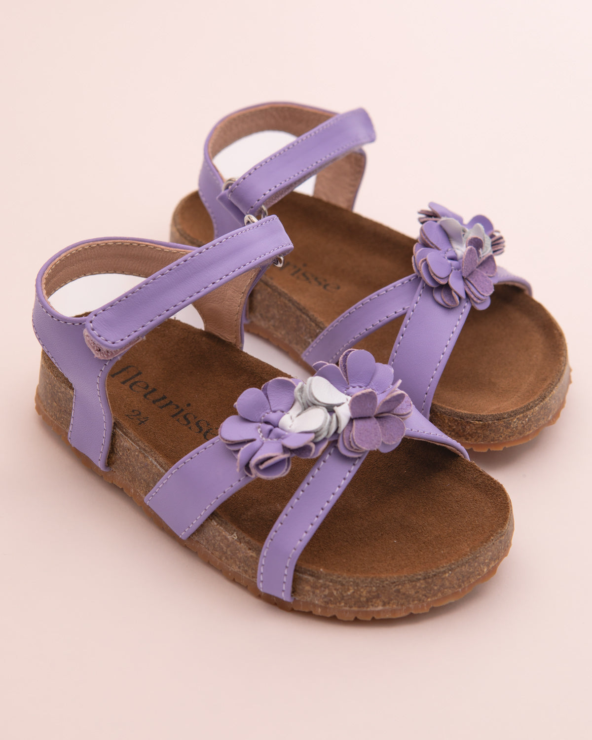 Pair of toddler lavender leather sandals with cork footbed and flower embellishments on the front straps, displayed on a blush pink background.