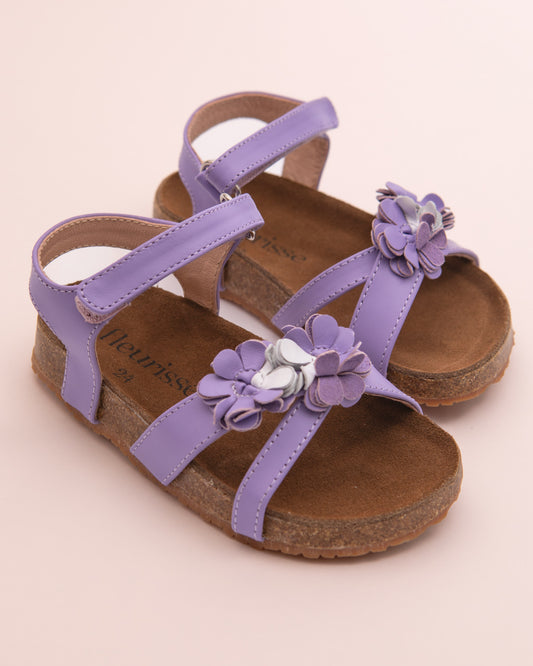 Pair of toddler lavender leather sandals with cork footbed and flower embellishments on the front straps, displayed on a blush pink background.