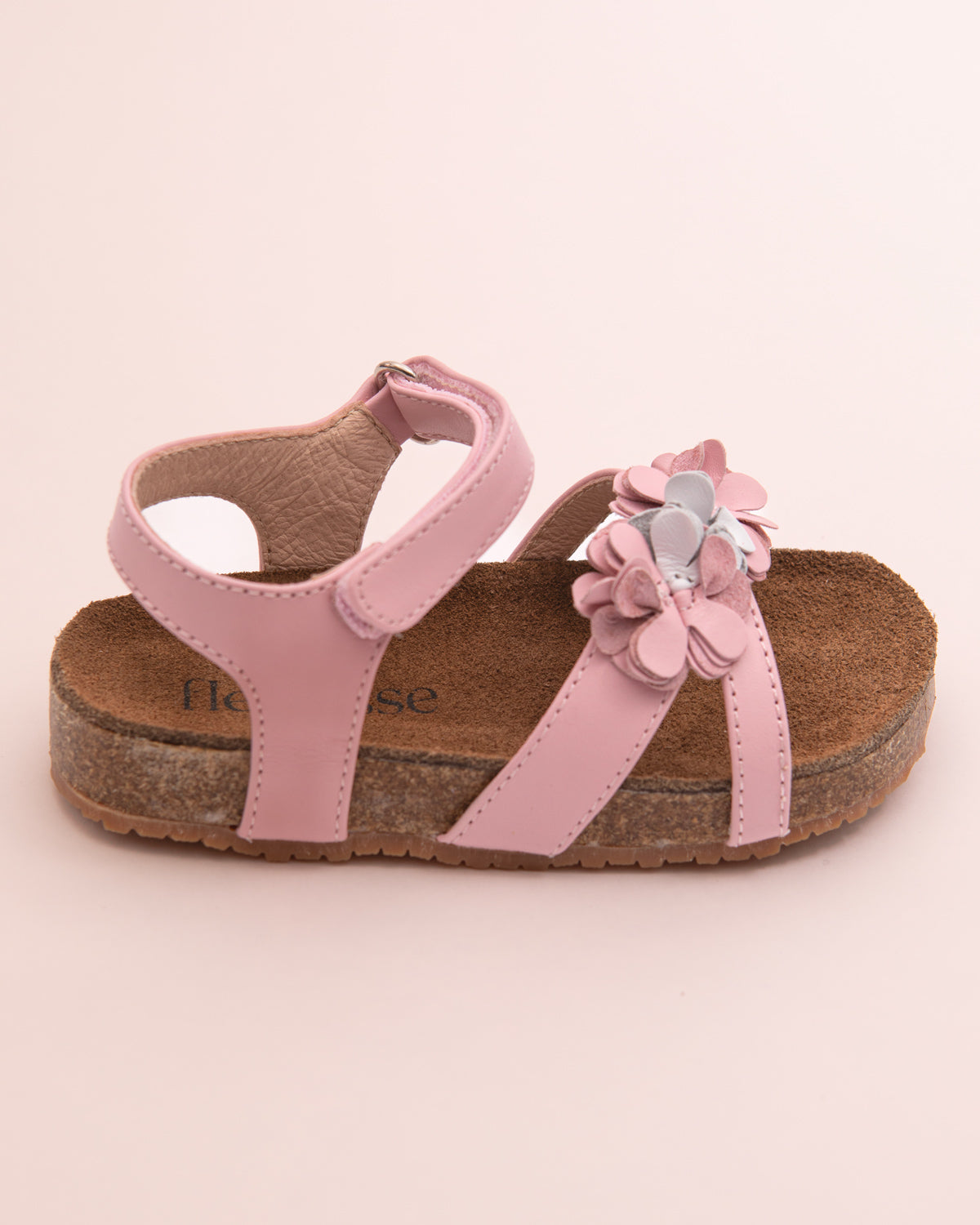 Side view of a blush pink leather toddler sandal with flower decorations and cork footbed, shown on a light pink surface.