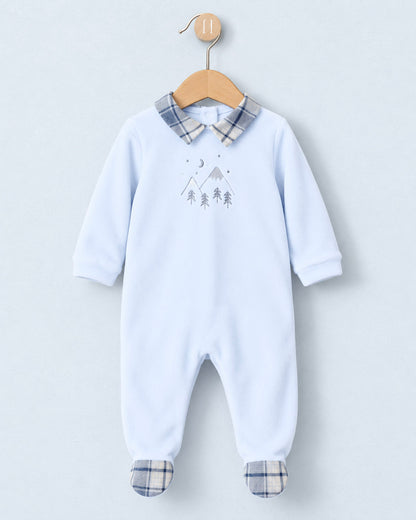 A baby footed one-piece jumpsuit in light blue, hanging on a wooden hanger against a light gray background. The jumpsuit features a plaid collar and matching plaid foot cuffs, with a small gray mountain and tree graphic on the chest.