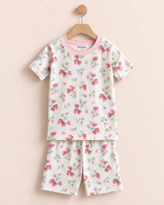 A two-piece children's pajama set hanging on a wooden hanger against a soft beige background. The set includes a short-sleeve top and matching shorts in an off-white fabric with an all-over pink and green floral print, finished with a solid pink crew neck.