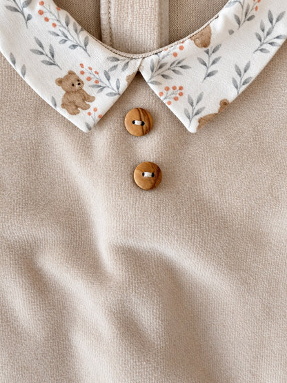 Close-up of a baby top featuring a solid beige knit body, a printed collar with a teddy bear and leaf pattern, and two wooden buttons.