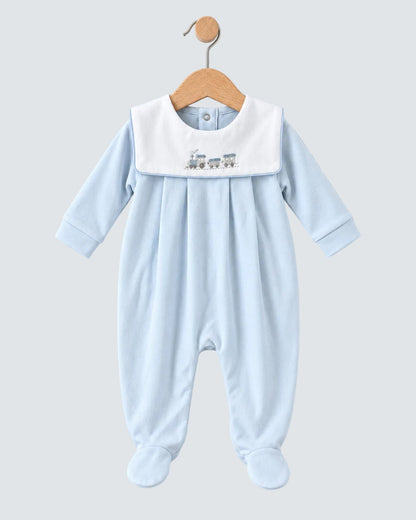 A baby footed sleeper in light blue with a contrasting white bib collar featuring a small embroidered train design, hanging on a wooden hanger against a light blue background.