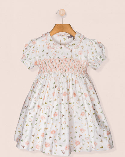 Full front view of the Valentina Soft Pink Toscana Smocked Dress displayed on a wooden hanger against a soft pink wall. The dress features puff sleeves, a floral print, and intricate smocking across the bodice.