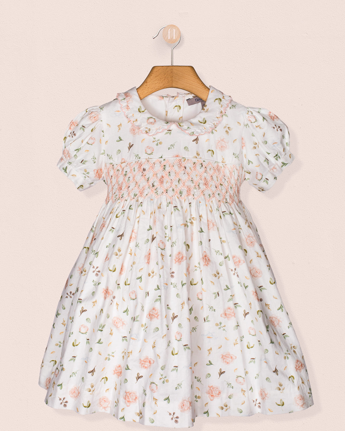 Full front view of the Valentina Soft Pink Toscana Smocked Dress displayed on a wooden hanger against a soft pink wall. The dress features puff sleeves, a floral print, and intricate smocking across the bodice.