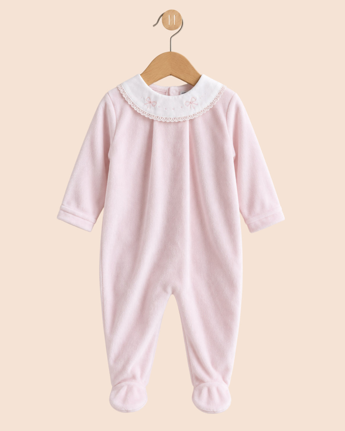 A light pink baby footed jumpsuit hanging on a wooden hanger against a solid beige background. The jumpsuit features a white embroidered collar with small bows and lace trim, and matching pink foot cuffs.
