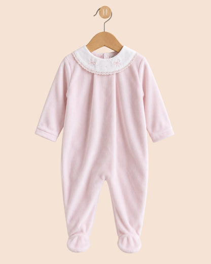 A light pink baby footed jumpsuit hanging on a wooden hanger against a solid beige background. The jumpsuit features a white embroidered collar with small bows and lace trim, and matching pink foot cuffs.