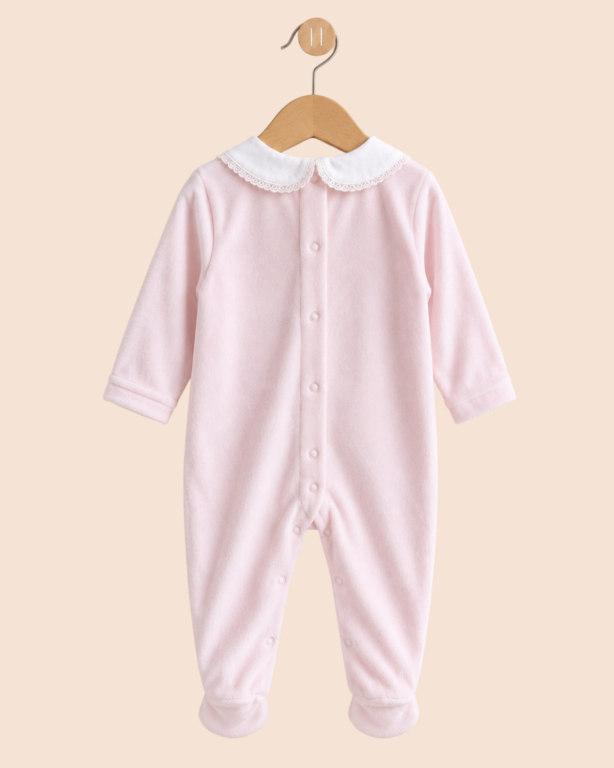 A light pink long-sleeve baby footed jumpsuit with a white Peter Pan collar, hanging on a wooden hanger against a solid beige background.