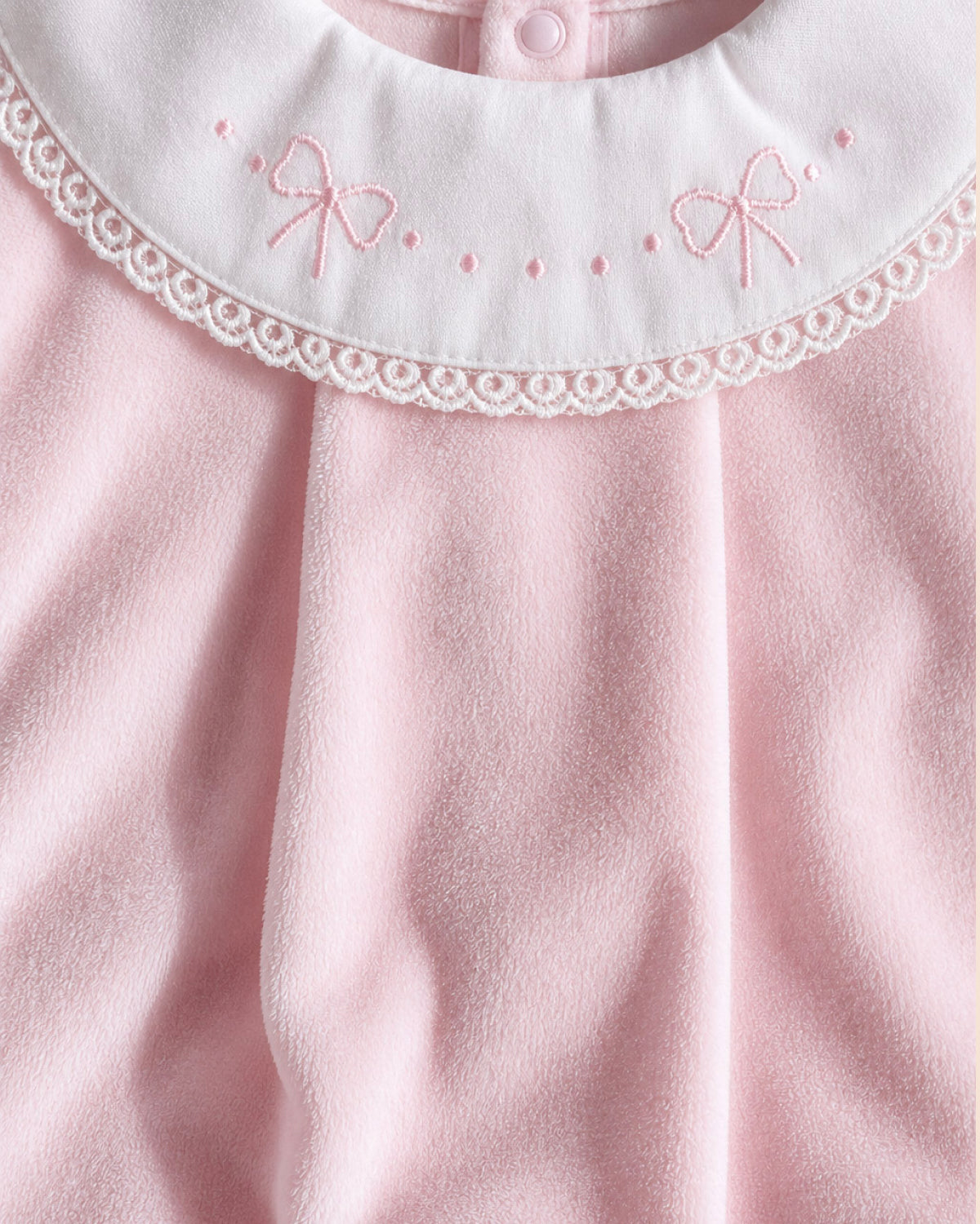 Close-up of a baby outfit featuring a soft pink fleece body and a contrasting white collar with pink embroidered bows and lace trim.