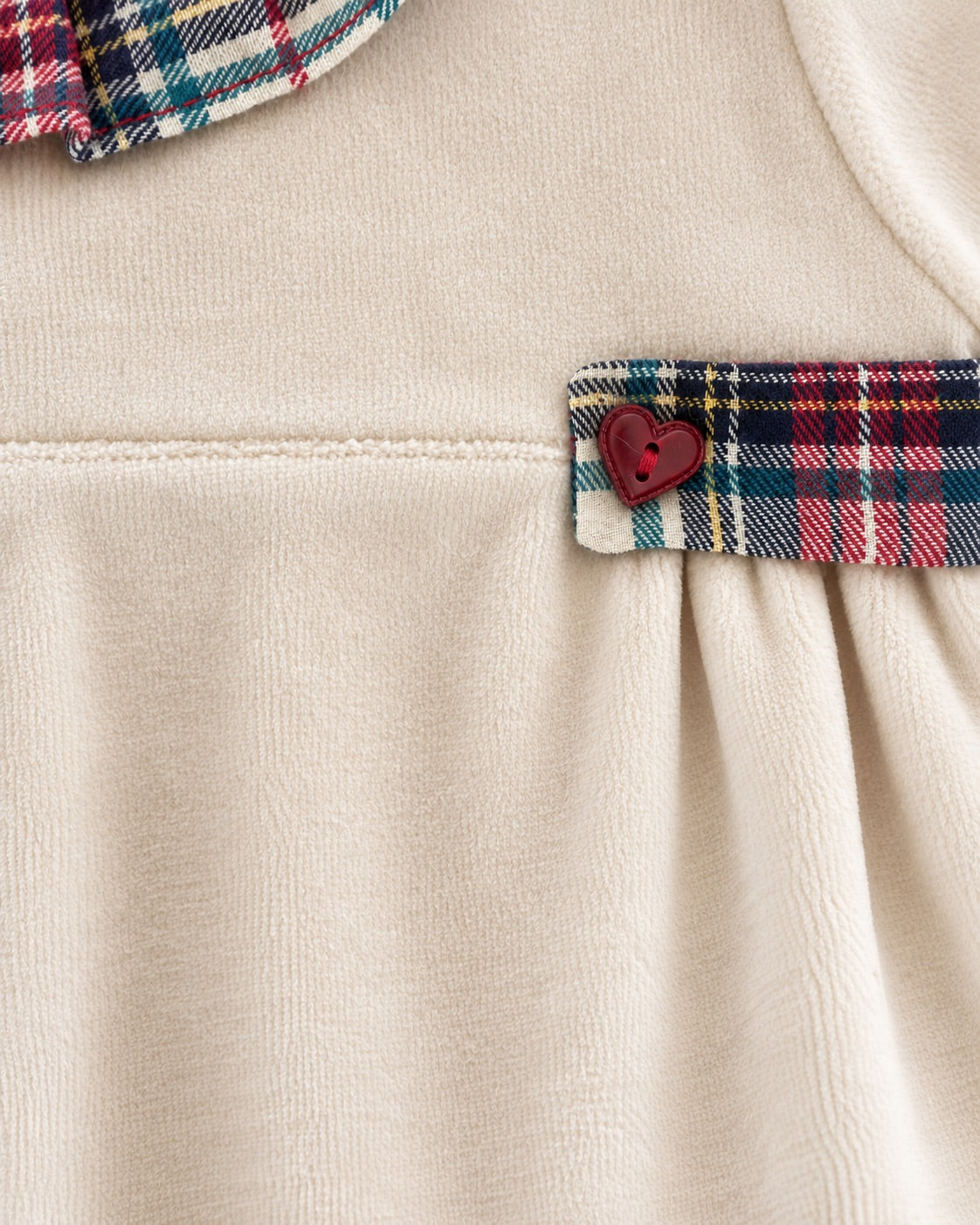 Close-up of a cream-colored garment featuring a red, blue, and white plaid accent band with a small red heart button, and part of a matching plaid collar at the top.