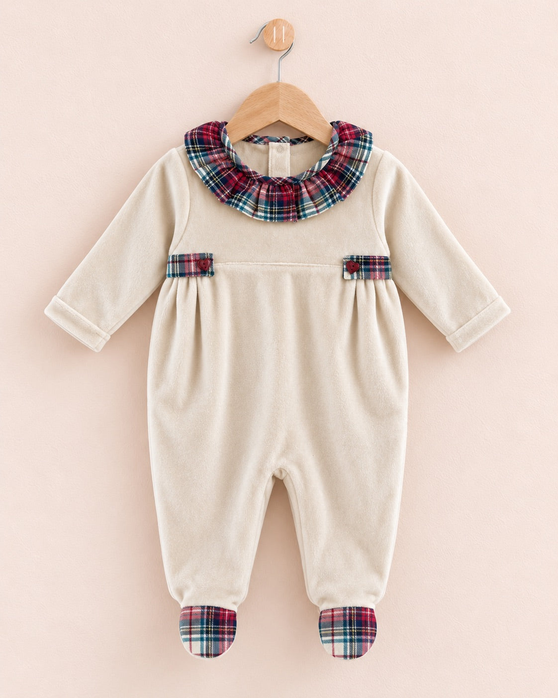 A cream-colored baby footed jumpsuit with a red, green, and blue plaid collar and matching plaid accents on the pockets and feet, hanging on a wooden hanger against a pale pink background.