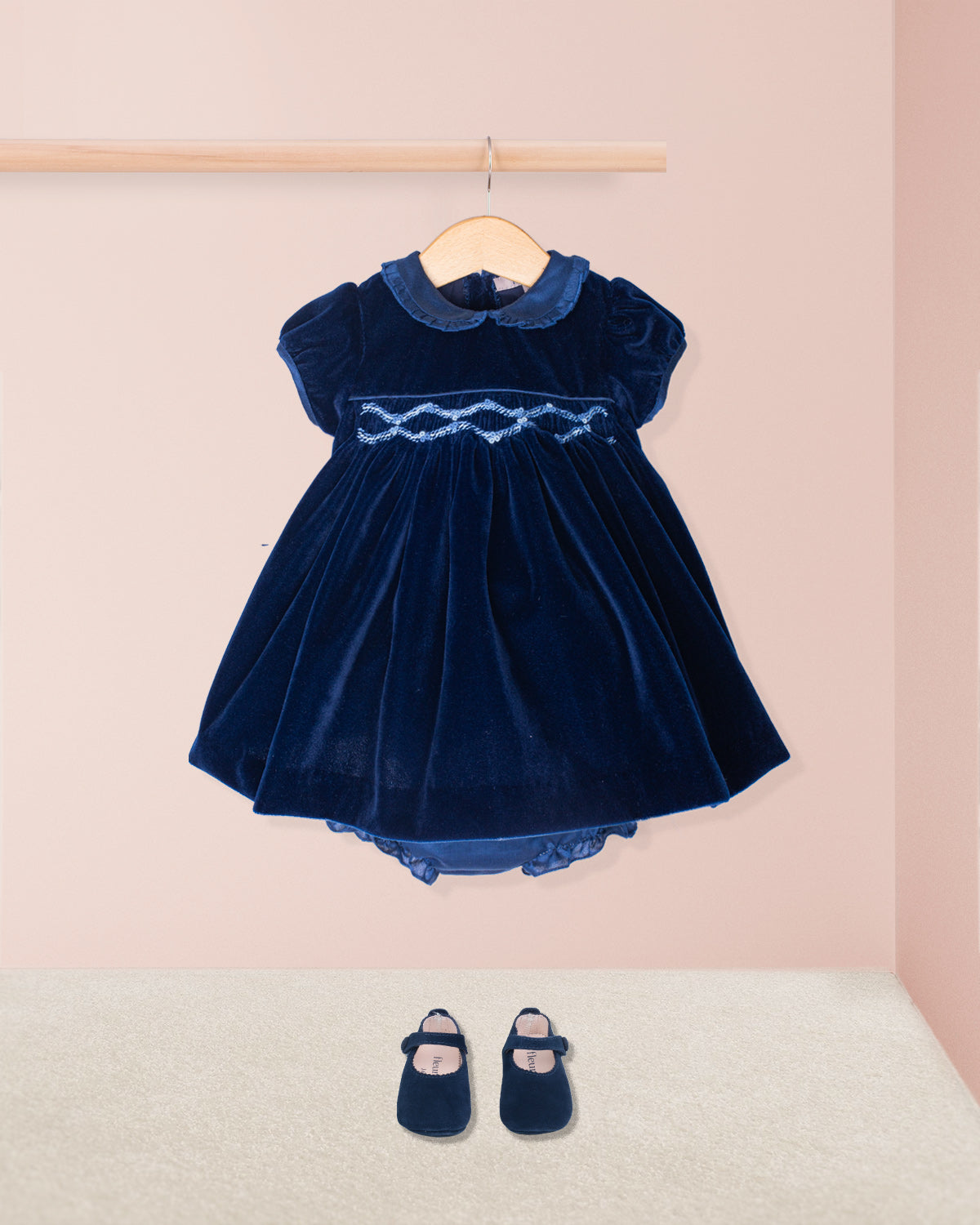 Navy blue velvet dress with lace details on a hanger against a pink wall.