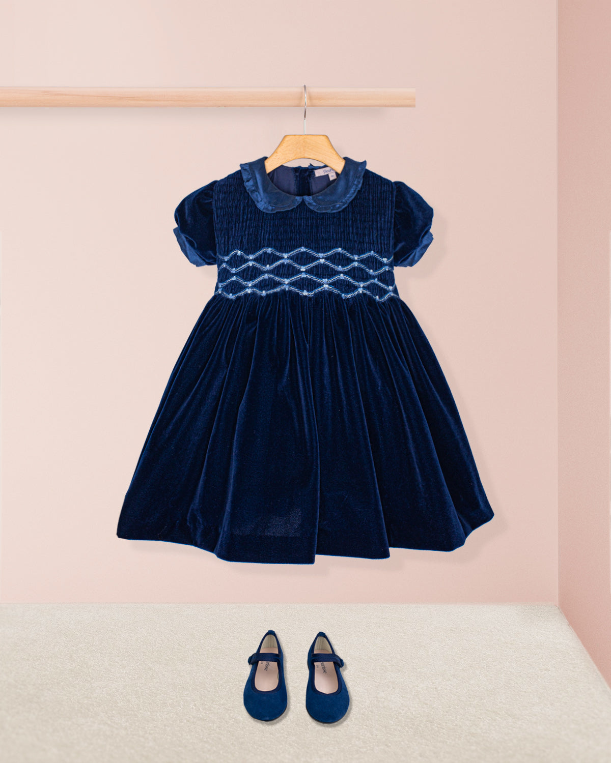 Navy blue velvet dress with white smocking on a pink background with shoes on hanger