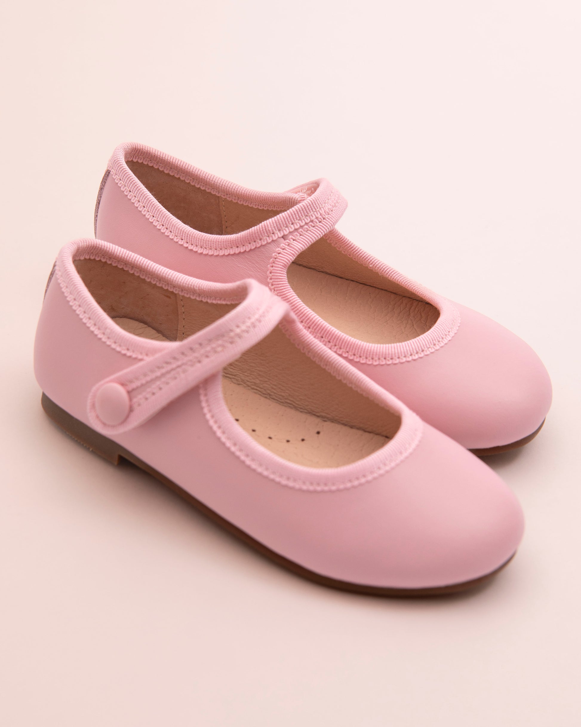 Pair of blush leather Mary-Jane shoes with matching velcro strap, grosgrain trim, and button detail, displayed on a soft pink background.
