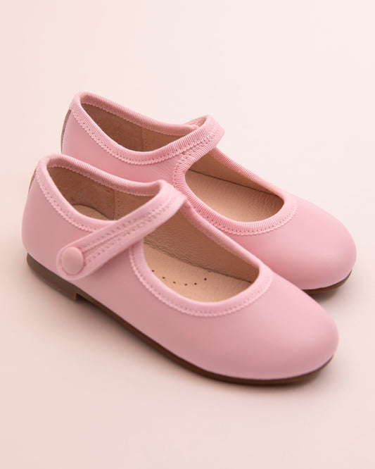 Pair of blush leather Mary-Jane shoes with matching velcro strap, grosgrain trim, and button detail, displayed on a soft pink background.