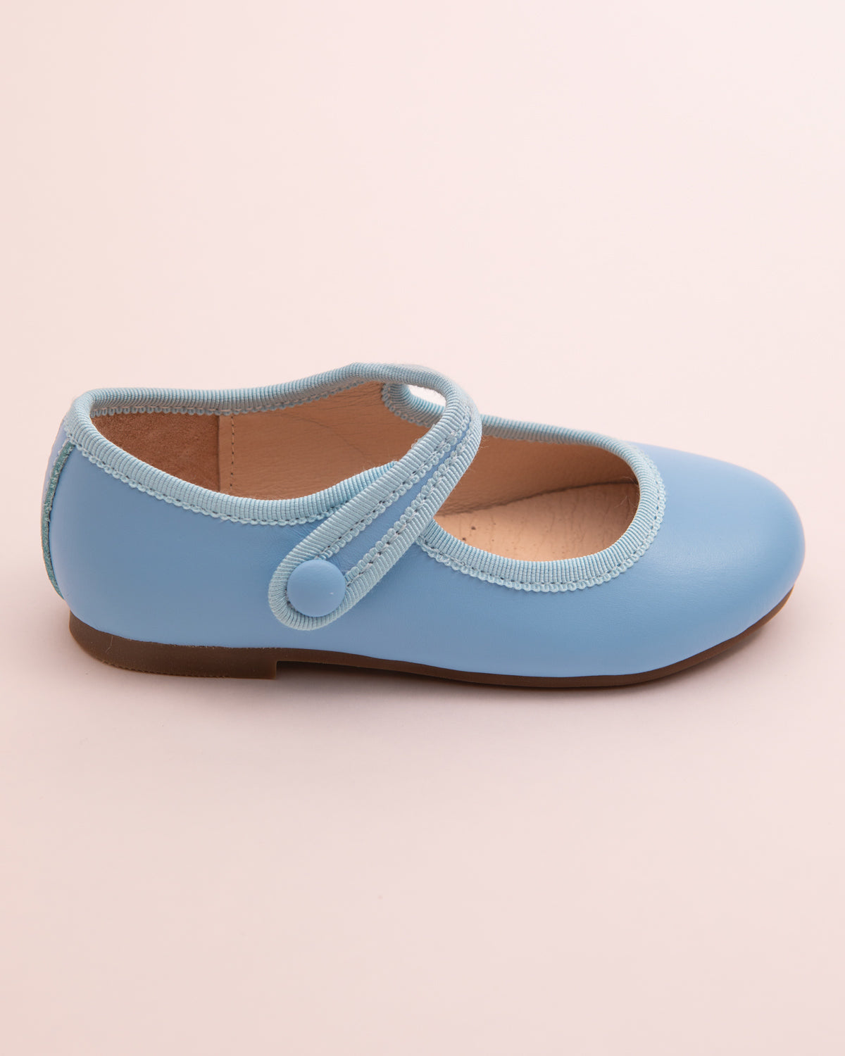 Side view of a single horizon blue leather Mary-Jane shoe with velcro strap, rubber sole, and tonal grosgrain trim on a light pink surface.