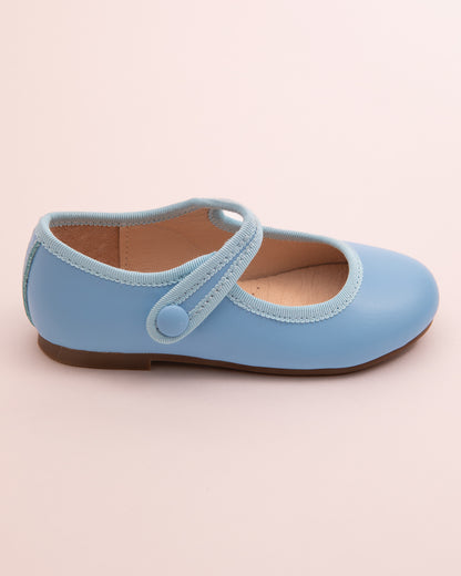 Side view of a single horizon blue leather Mary-Jane shoe with velcro strap, rubber sole, and tonal grosgrain trim on a light pink surface.