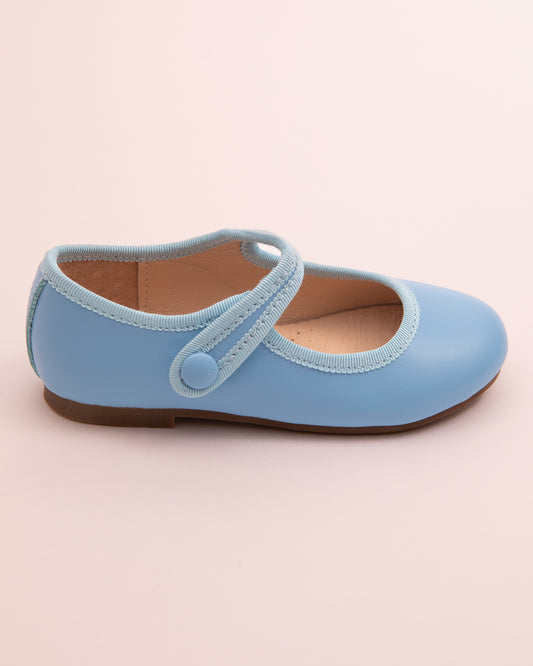 Side view of a single horizon blue leather Mary-Jane shoe with velcro strap, rubber sole, and tonal grosgrain trim on a light pink surface.