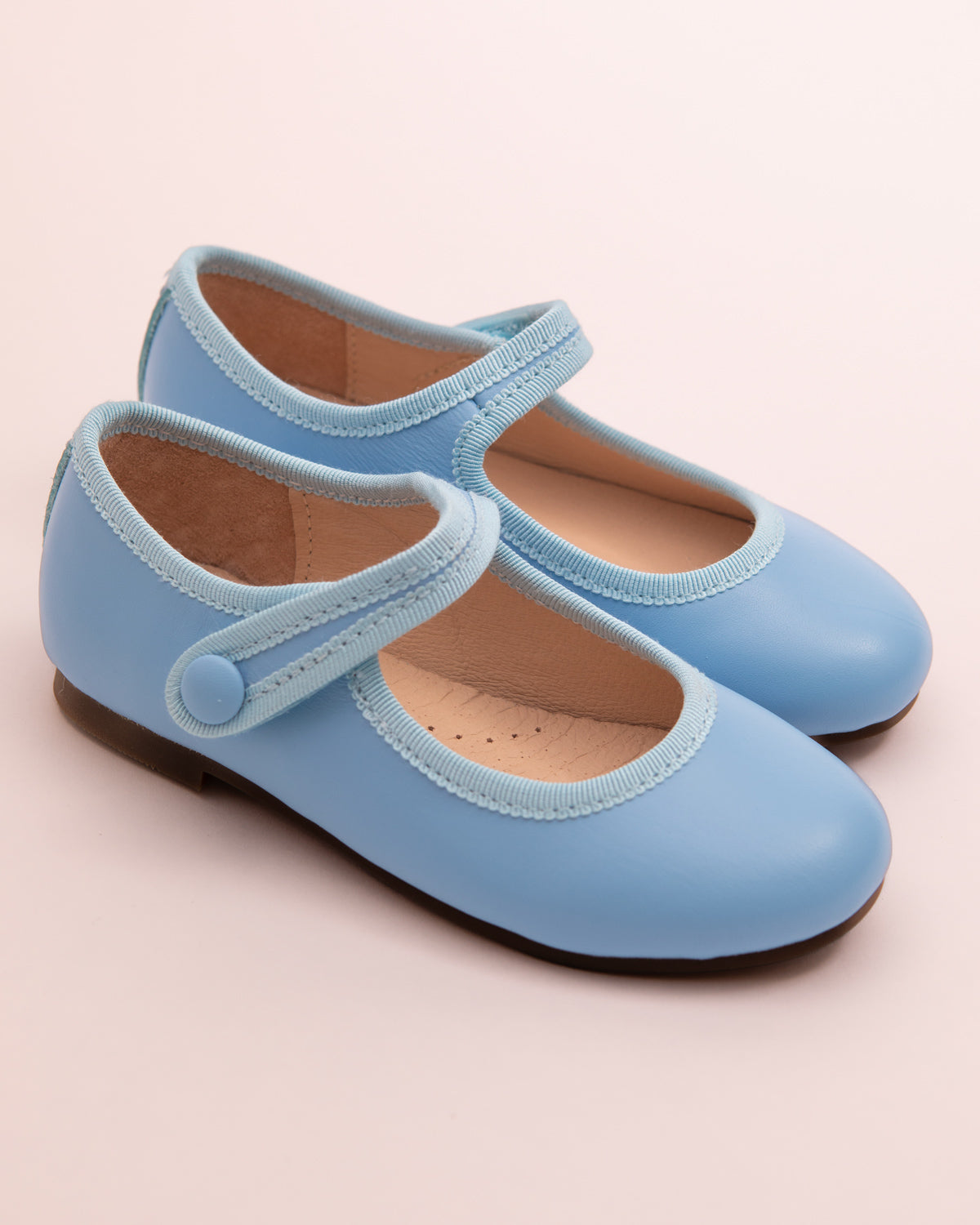 Pair of horizon blue leather Mary-Jane shoes with matching velcro strap, grosgrain trim, and button detail, displayed on a soft pink background.