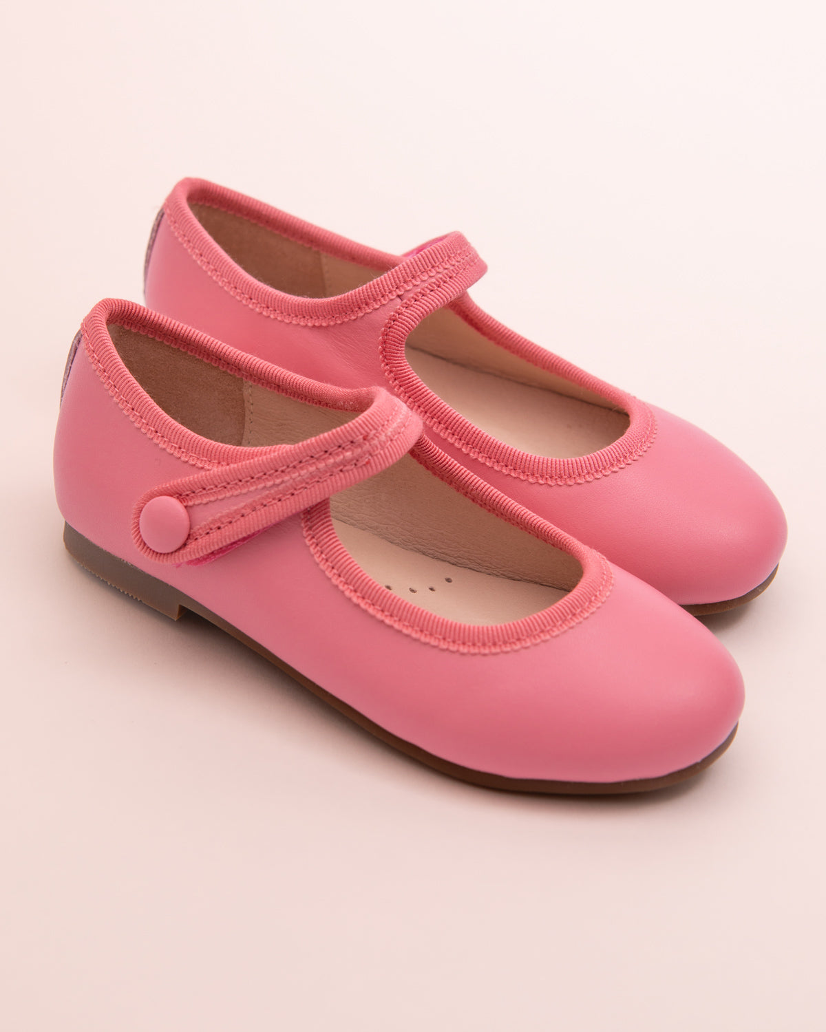 Pair of primrose leather Mary-Jane shoes with matching velcro strap, grosgrain trim, and button detail, displayed on a soft pink background.