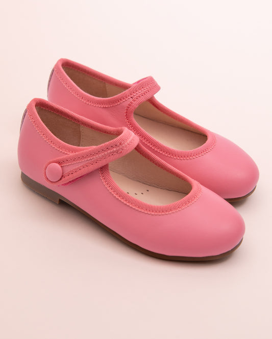 Pair of primrose leather Mary-Jane shoes with matching velcro strap, grosgrain trim, and button detail, displayed on a soft pink background.