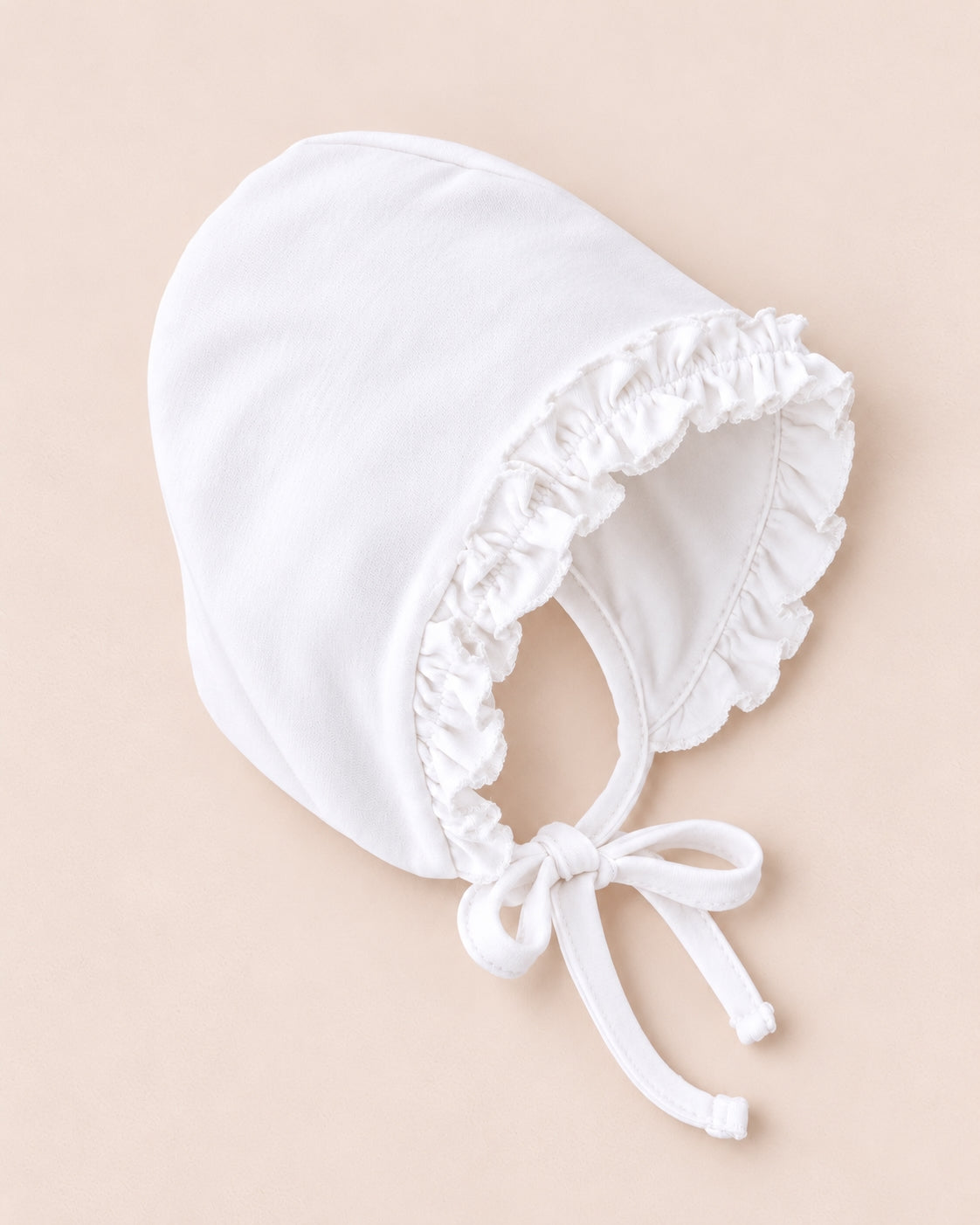 A white bonnet-style baby hat with ruffled trim and tie strings, photographed on a soft beige background.