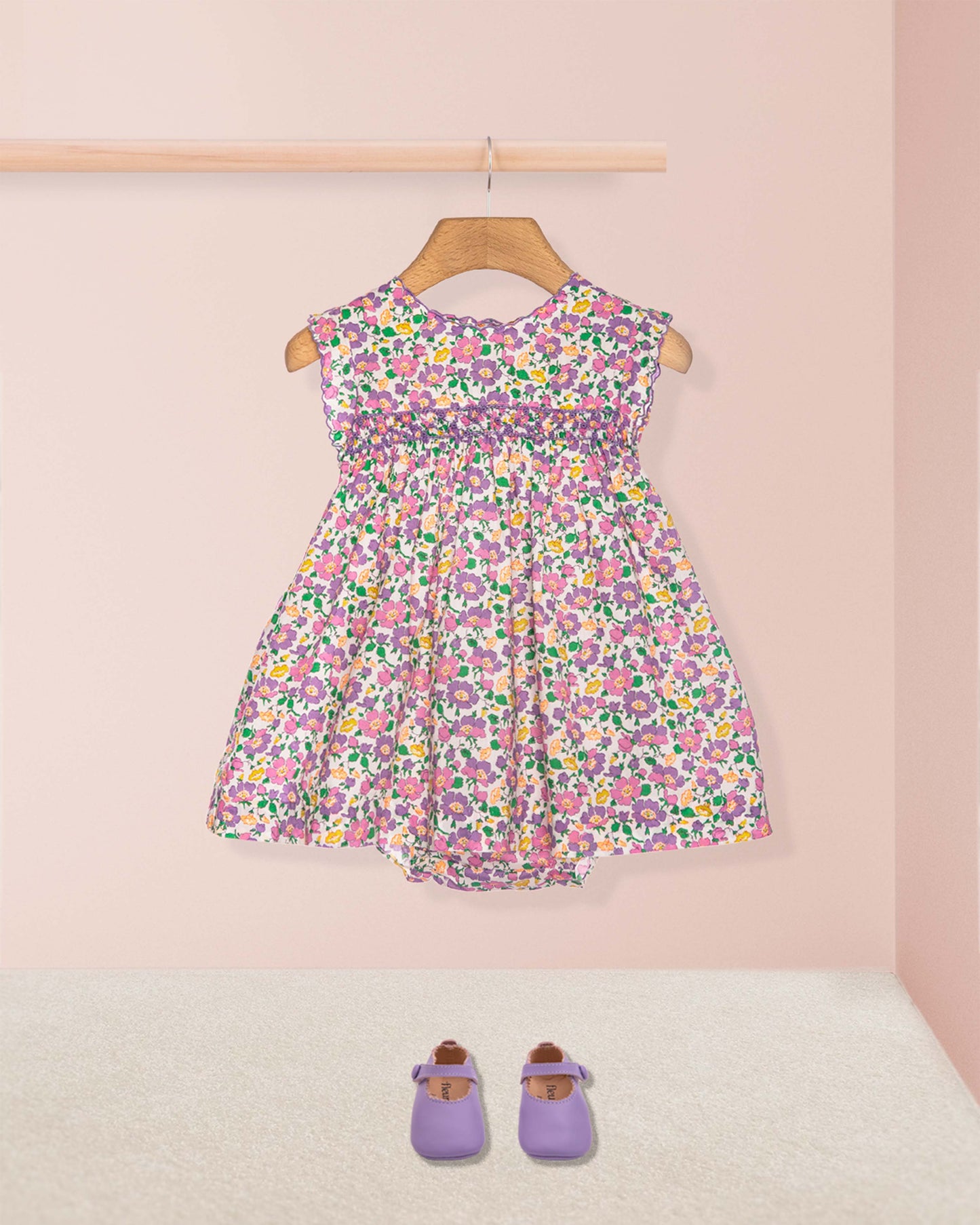 Floral dress on a hanger with purple shoes on the floor against a pink wall.