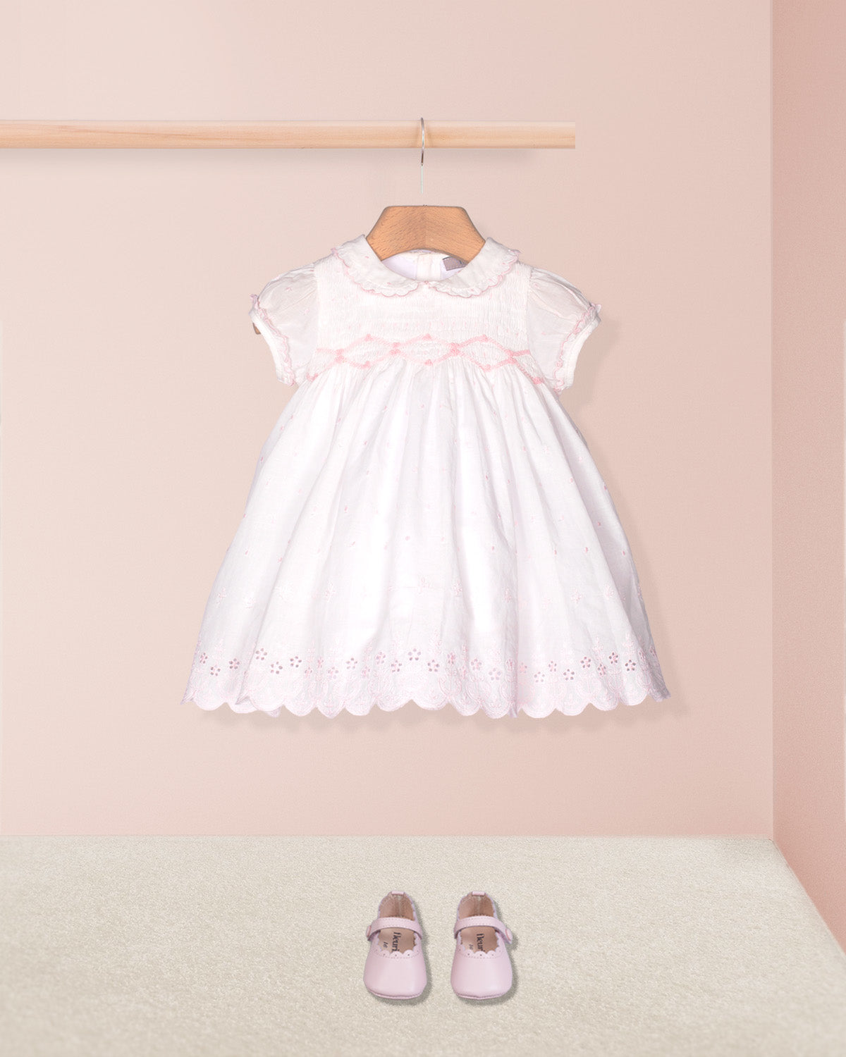 White dress with pink trim on a hanger against a light pink wall, with matching shoes on the floor.