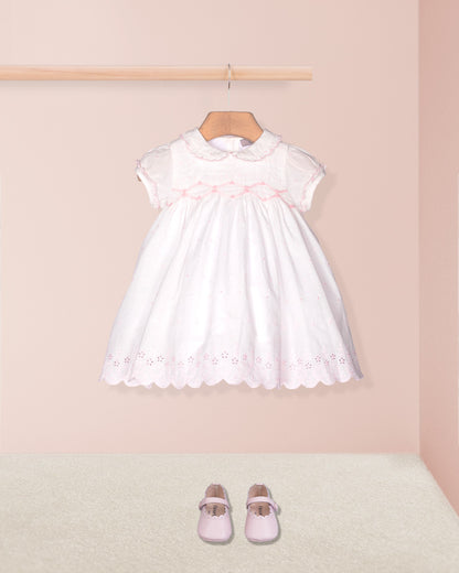 White dress with pink trim on a hanger against a light pink wall, with matching shoes on the floor.